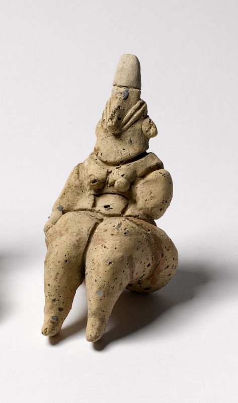 A small clay figurine of a seated figure with a pointed head, slit 'coffee-bean' eyes, and broad hips, modeled in pale fired clay.