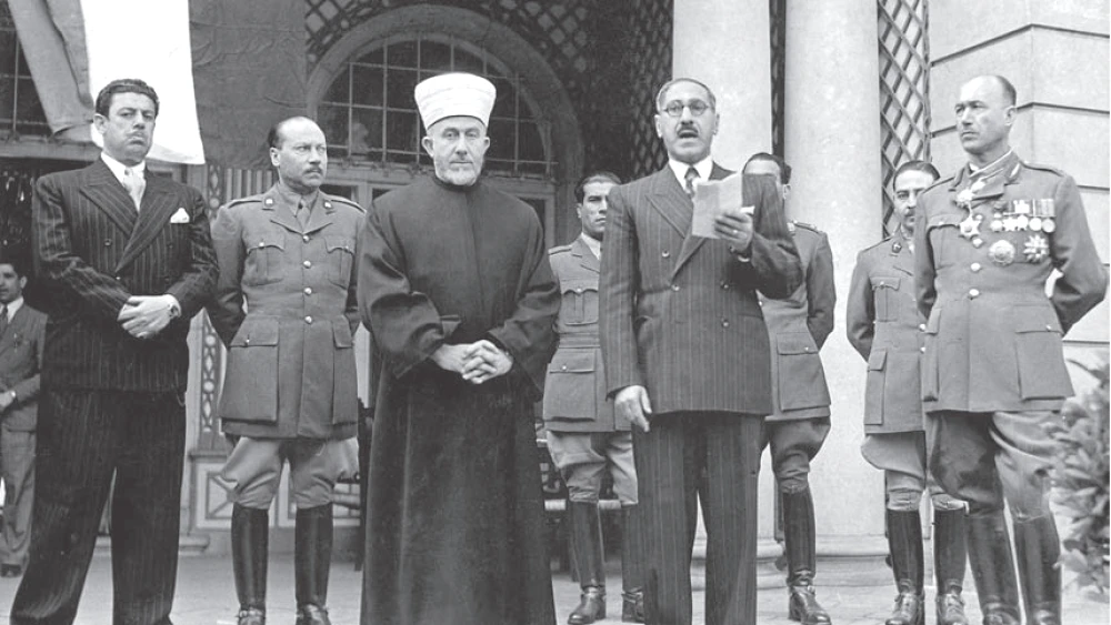 A black-and-white photograph outside an arched building: Yunis Bahri, in glasses and a dark suit, reads from a paper beside Amin al-Husseini, in white turban and robe, flanked by men in German military uniforms.
